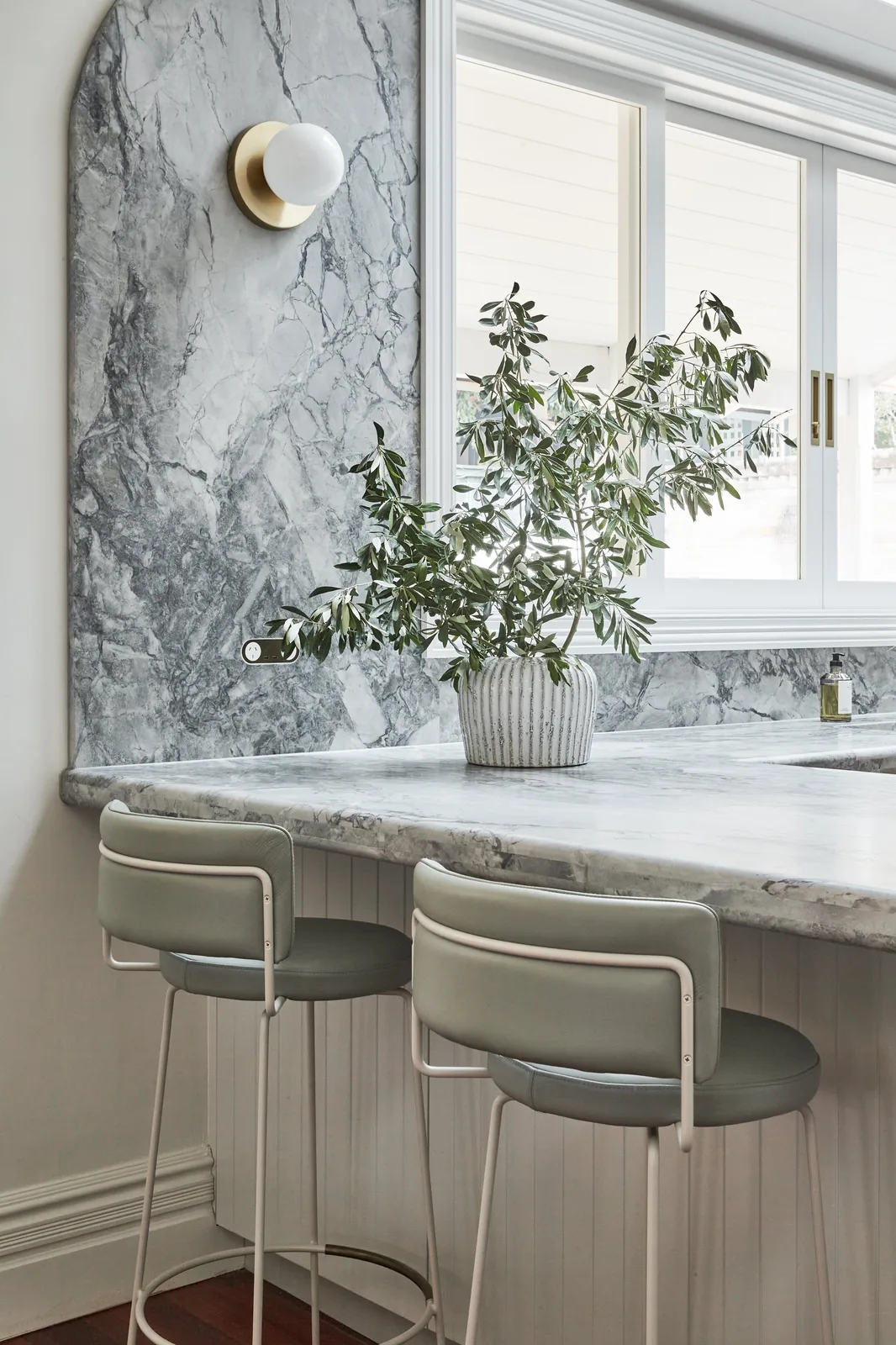 Elegant kitchen bench with seating, marble benchtop and splashbackwith soft natural light from windows shining on a small potted plant.