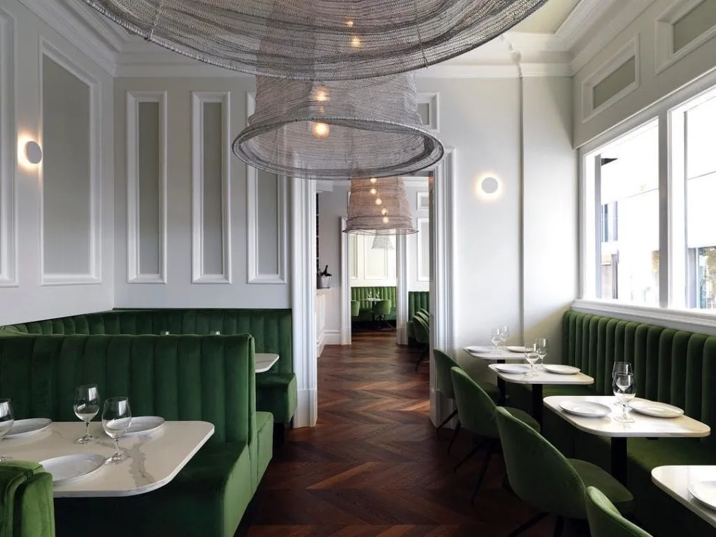 An art deco style restaurant seating area with dark hardwood flooring, light walls featuring decorative wainscoting, emerald green booths and seating. Tables are laid with plates and wine glasses ready for diners.