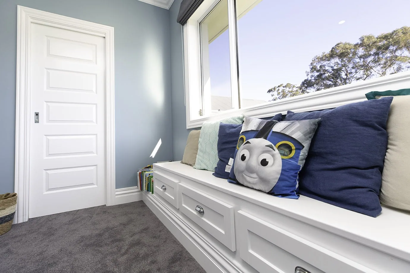 A boy's bedroom with bench seat underneath a window, painted white, and Intrim SK460 skirting and architrave framing a white door. Grey carpet lines the floors and a Thomas the Tank Engine pillow sits on the bench seat.