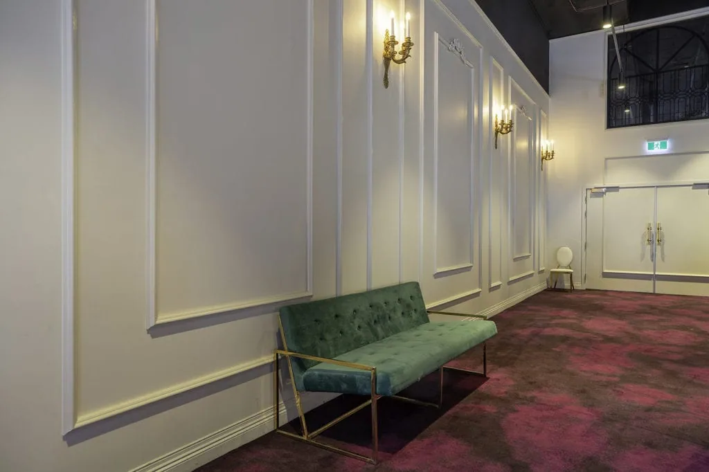 1920s-style waiting area with white panelled walls, red patterned carpet, and a green sofa with gold detailing