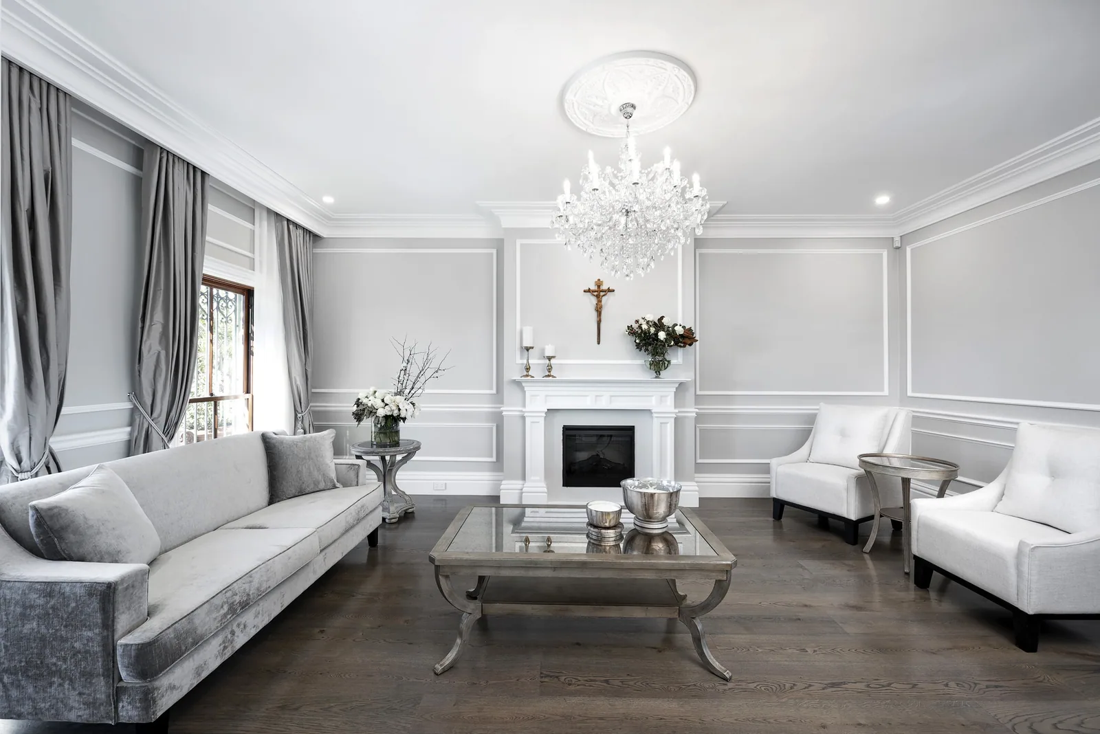 Elegant grey and white living space in 1920s style adorned with plush furnishings, a glass accent table, and ornate chandelier