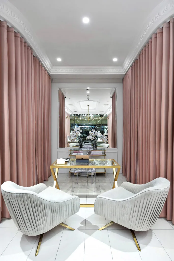 A plush space in a retail shop for customers to sit. A white tile floor is surrounded on both sides with pale pink curtains and the back wall features floor to ceiling wainscoting with a large decorative mirror reflecting back into the rest of the shop.