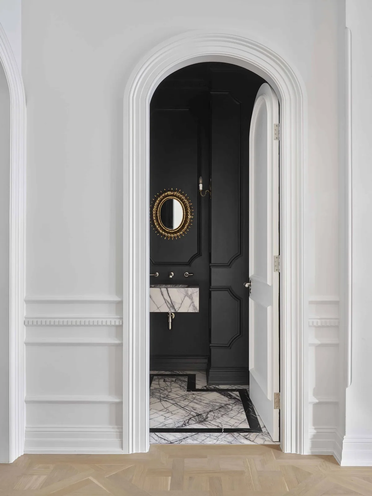 Corridor and powder room at Matchpoint House Strathfield by Duet, viewed through a white arched opening with Intrim custom curved architraves, CR84 dado rail and SK173 skirting, leading to a dramatic black-painted powder room with gold sunburst mirror and marble vanity